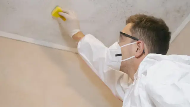 Worker in a mask and protective suit wiping a stained ceiling corner